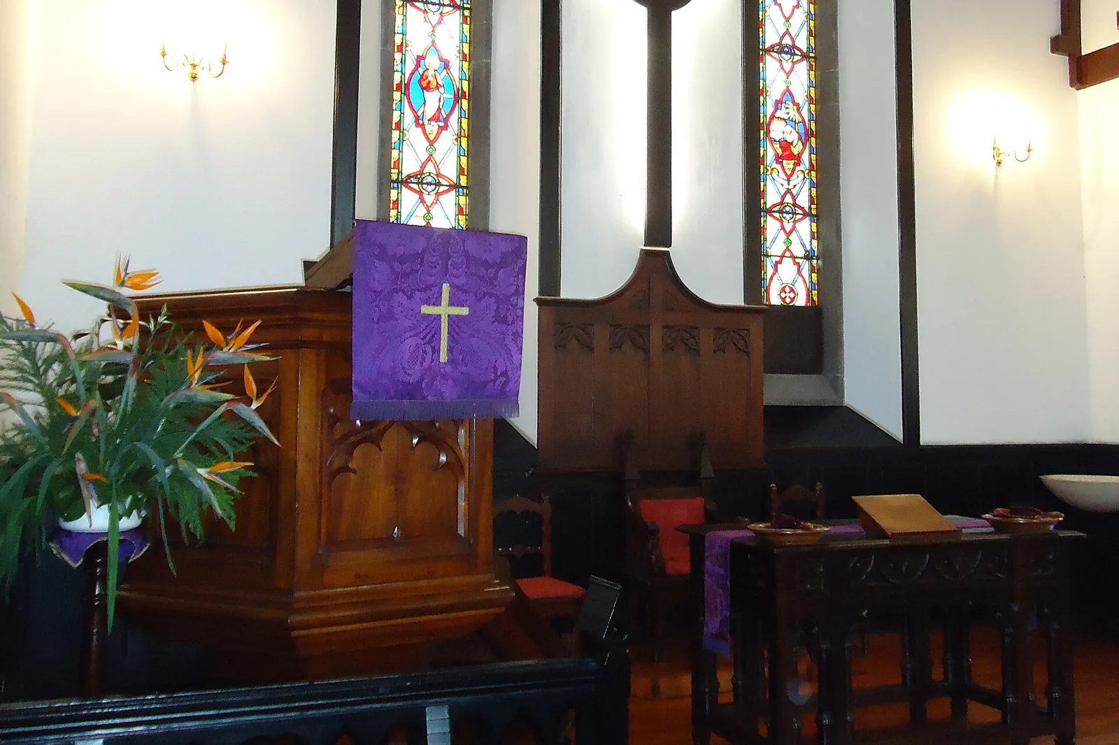 The chancel of the church with a wooden pulpit, a purple cloth, a cross and two stained-glass windows in pointed arches
