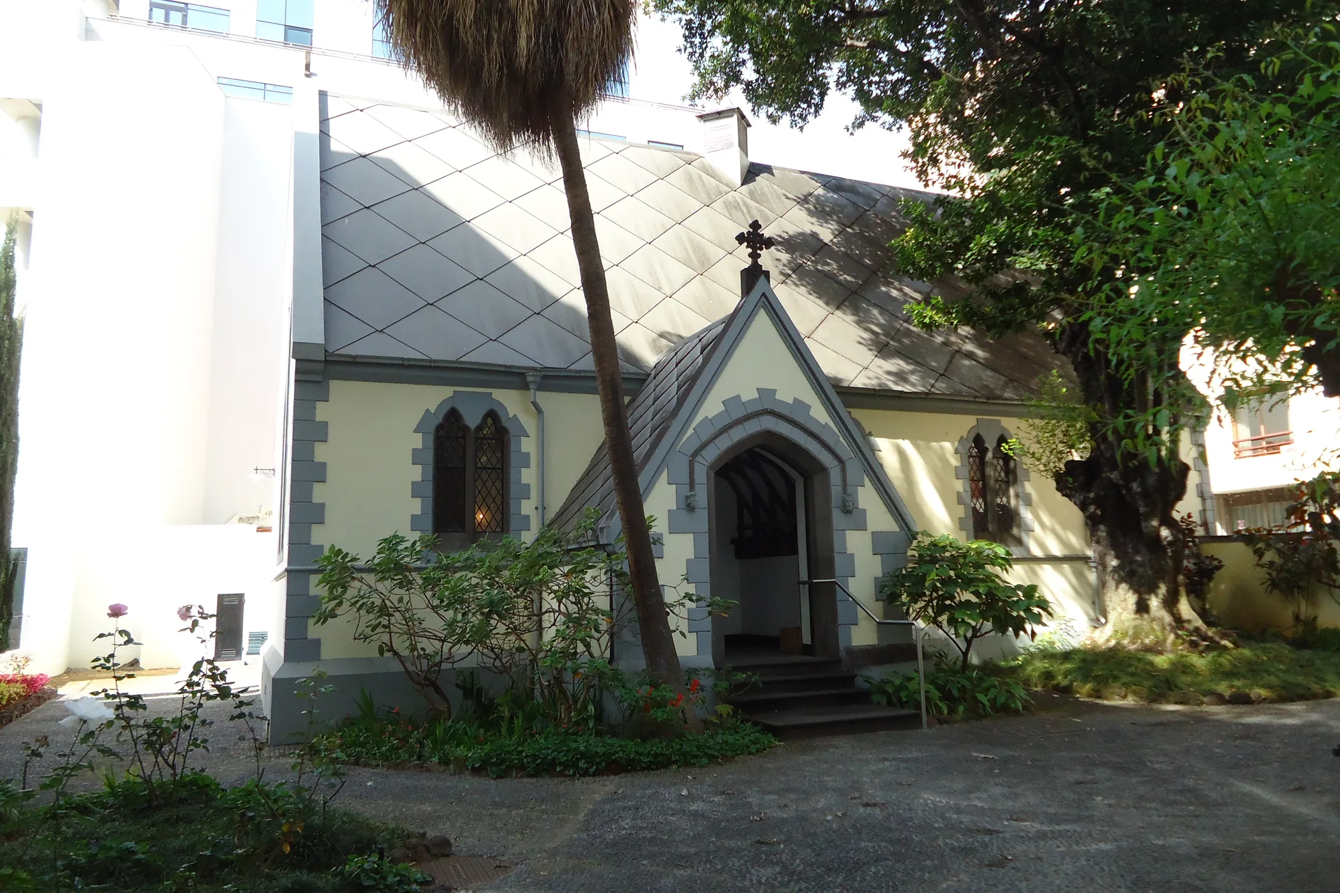 St Andrew's Presbyterian church in Funchal, a small neo-Gothic church with a grey diamond-patterned roof, pale yellow walls, grey stone trim and a pointed arched doorway, seen across a garden with a tall palm leaning in front